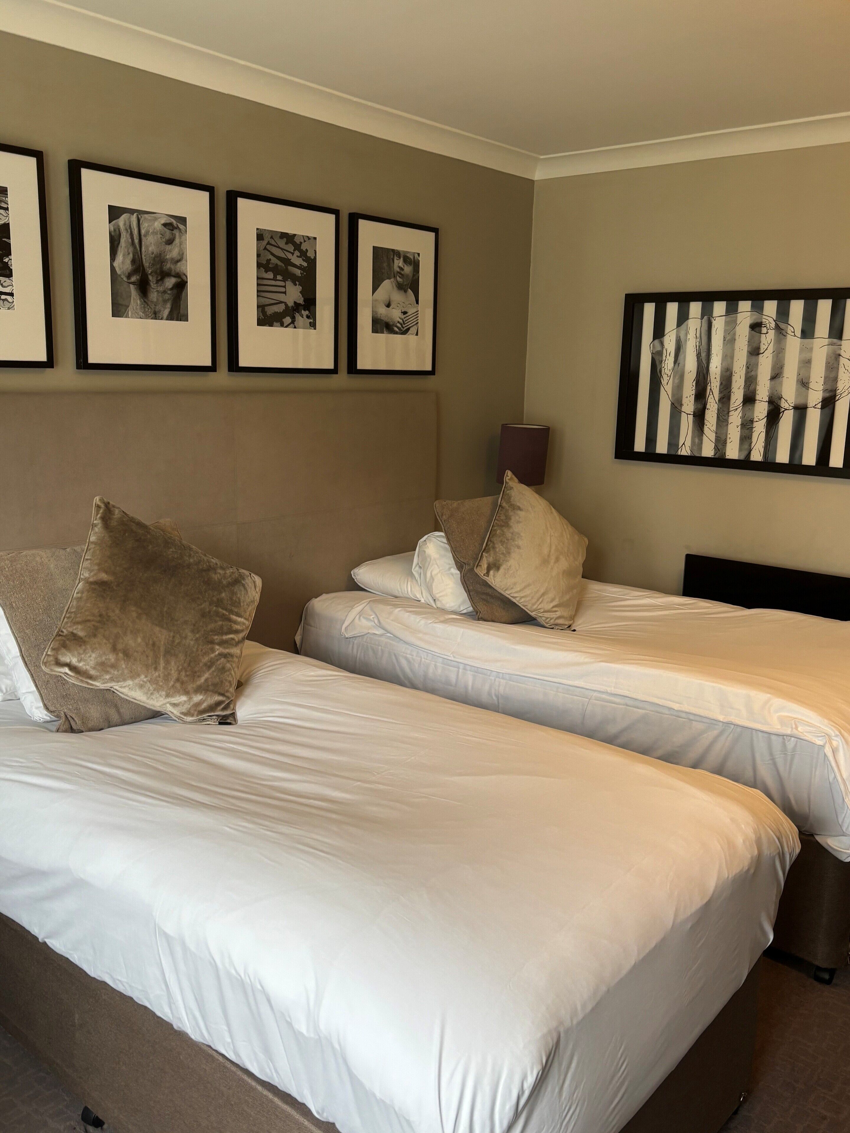 A hotel room with two beds, each with white linens and multiple pillows. There are framed pictures on the wall above the beds.