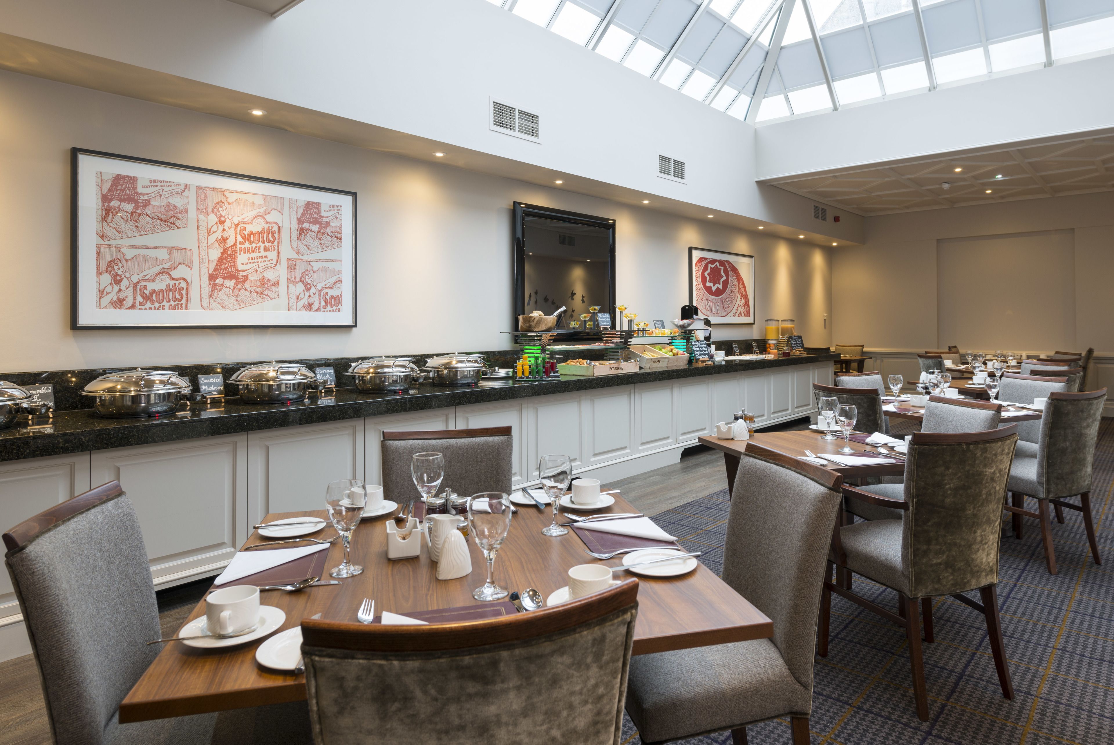 A hotel dining area with a high ceiling and skylight, featuring a buffet with various food items and a seating area with tables set for dining.