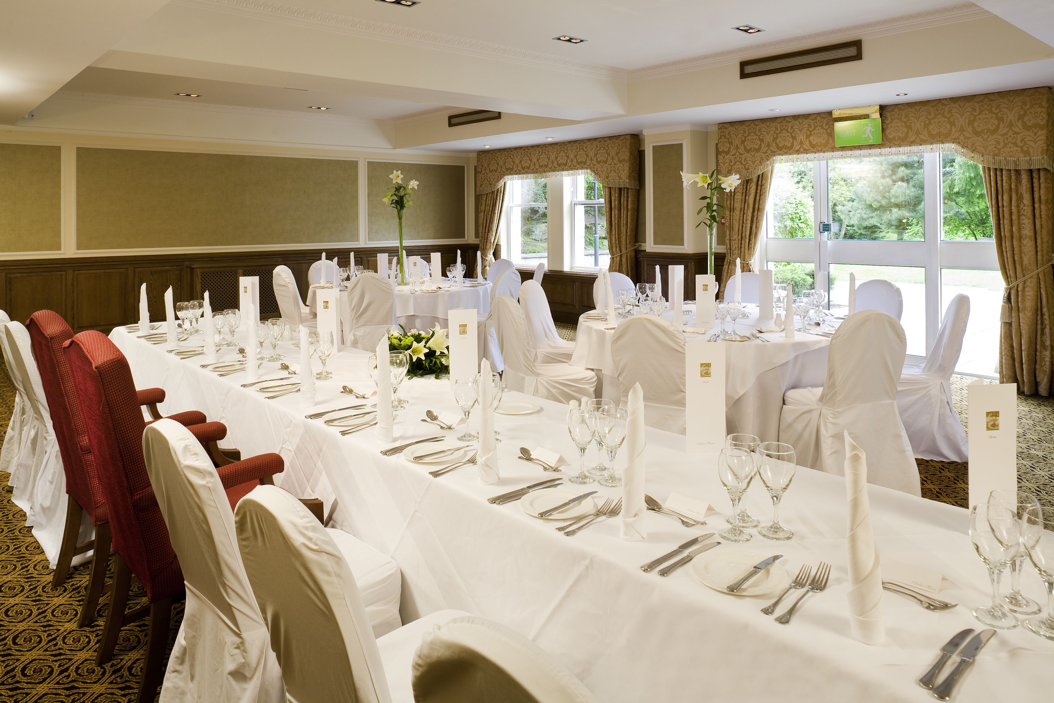 A banquet hall with long tables set for dining, white chairs, and a view of greenery outside.