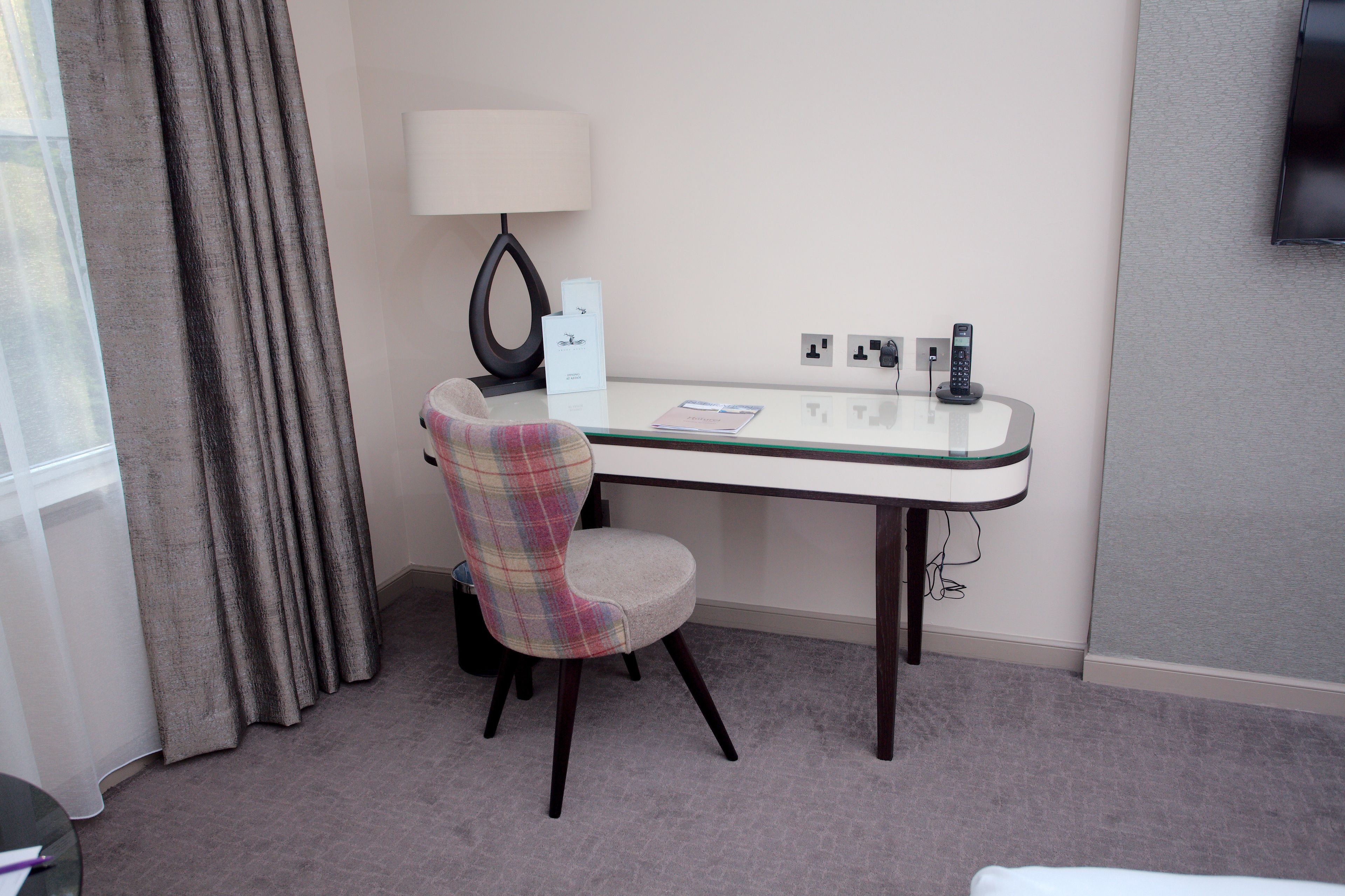 A hotel room with a desk, a chair, a lamp, a telephone, and a TV mounted on the wall.