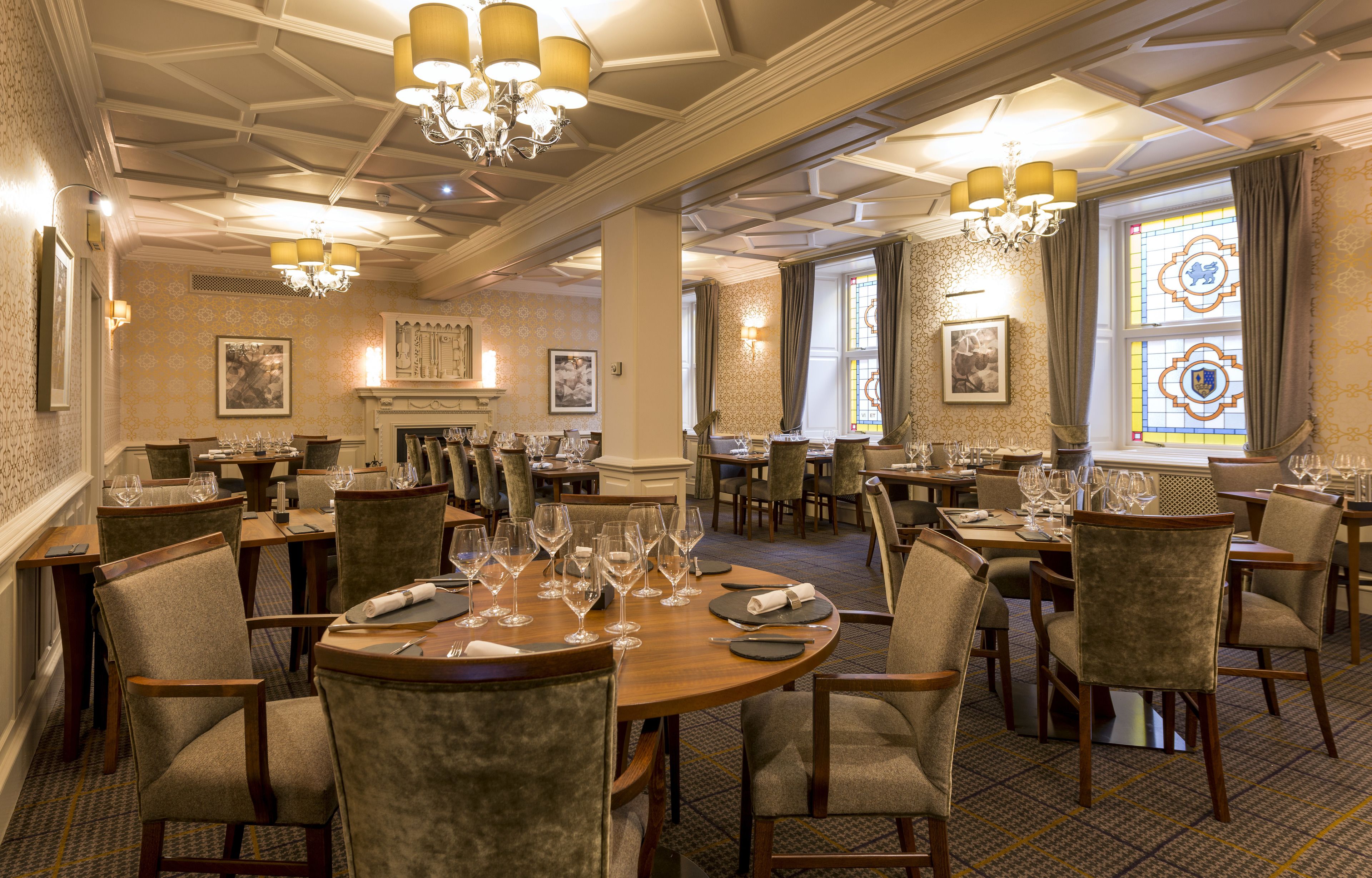 A formal dining room with multiple tables and chairs, chandeliers, and patterned wallpaper.