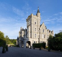 Um edifício grande e histórico de pedra, com várias torres e portões em arco, rodeado por um jardim bem cuidado e caminhos pavimentados.