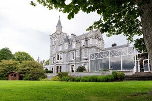 Un edificio grande y histórico de piedra con una torre destacada, un invernadero y un césped bien cuidado.