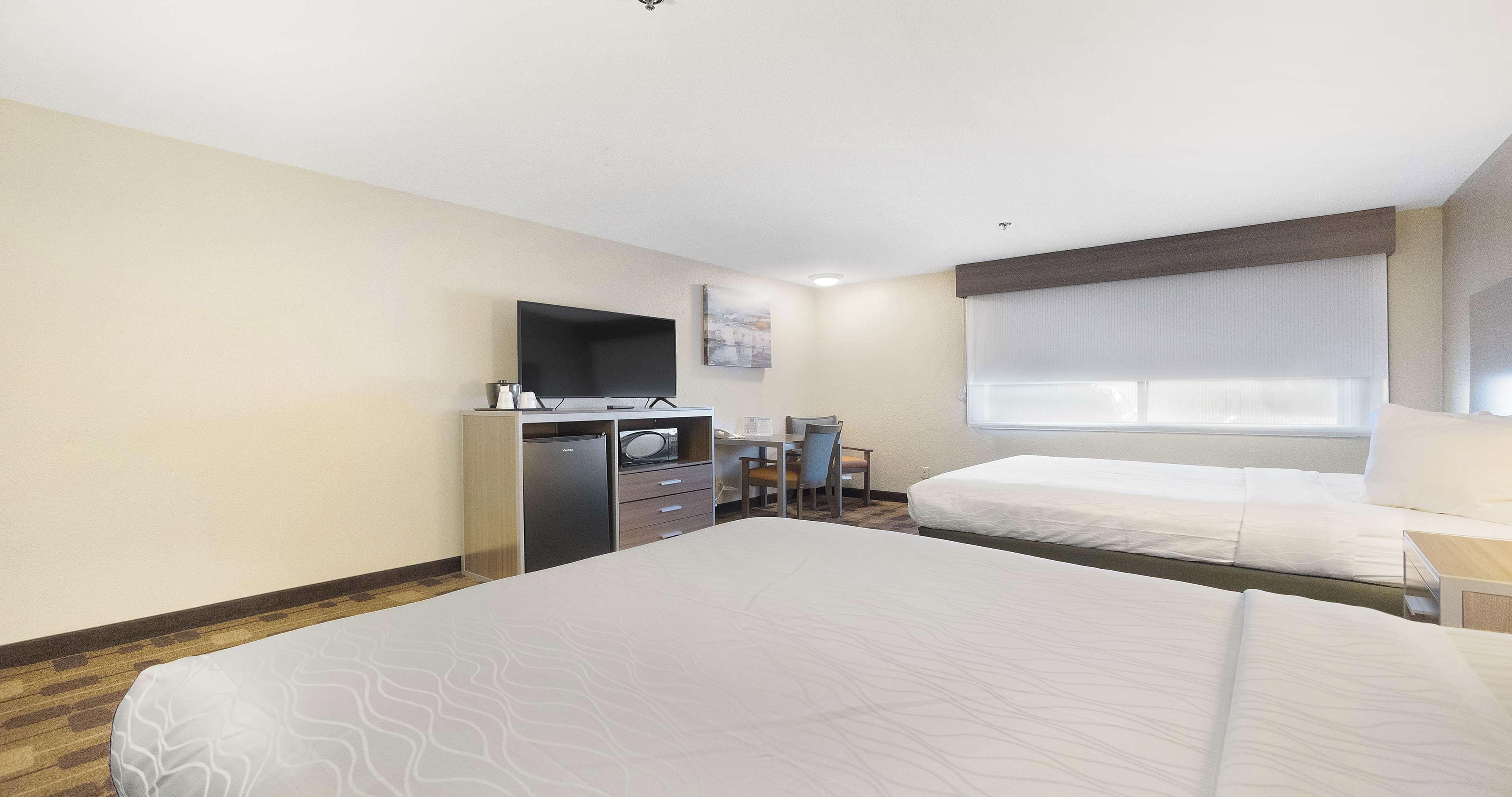 A hotel room with a large bed, a desk with a flat-screen TV, a small table, and a window with blinds.