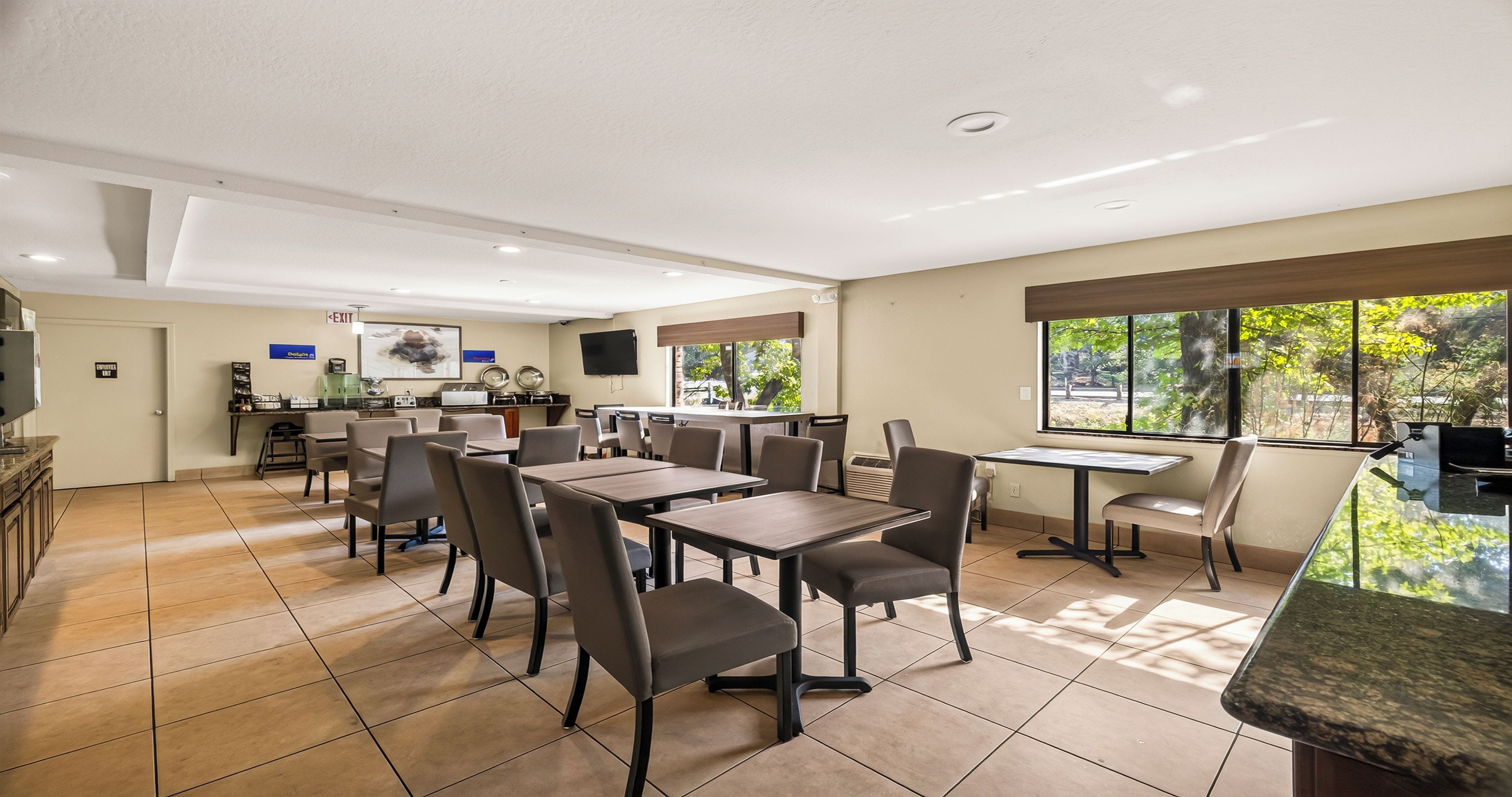 A hotel dining area with multiple tables and chairs, a kitchen in the background, and a view of the outdoors through large windows.