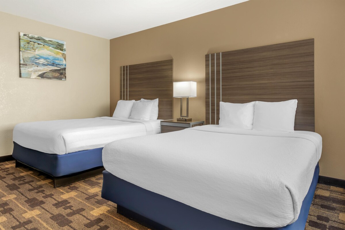 A hotel room with two beds, a nightstand, a lamp, and a painting on the wall.