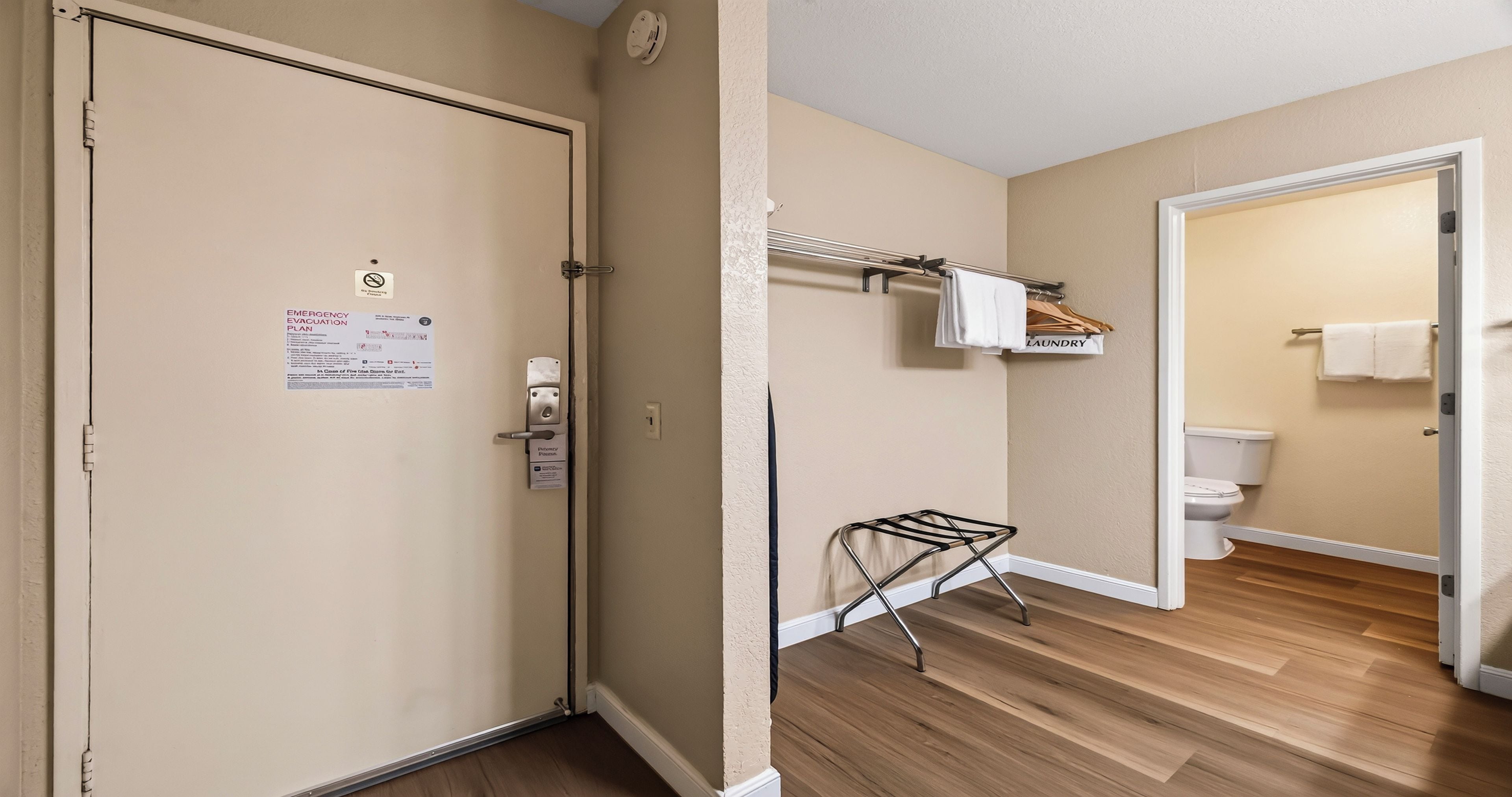 Una camera d'albergo con pavimento in legno, una porta con il piano di evacuazione di emergenza, un angolo lavanderia con un'asta appendiabiti e un water.