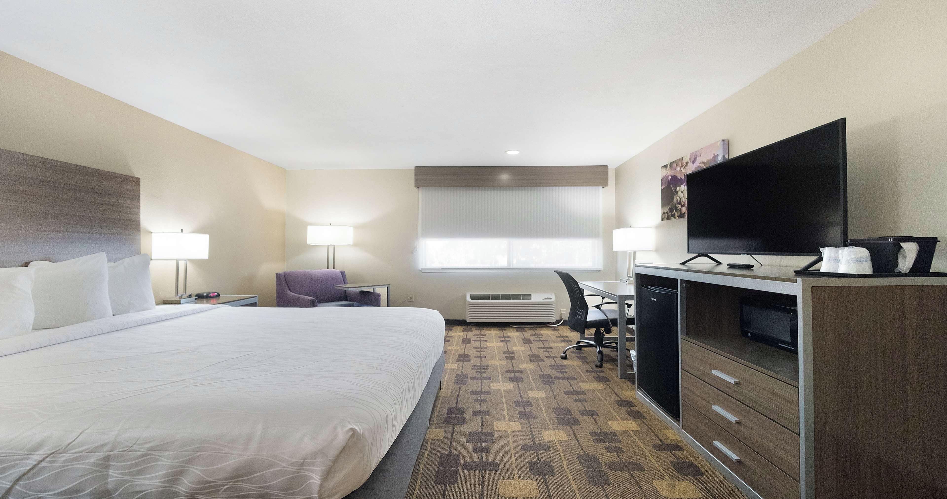 Een hotelkamer met een groot bed, een bureau met computer, een flatscreen televisie en een raam met jaloezieën.
