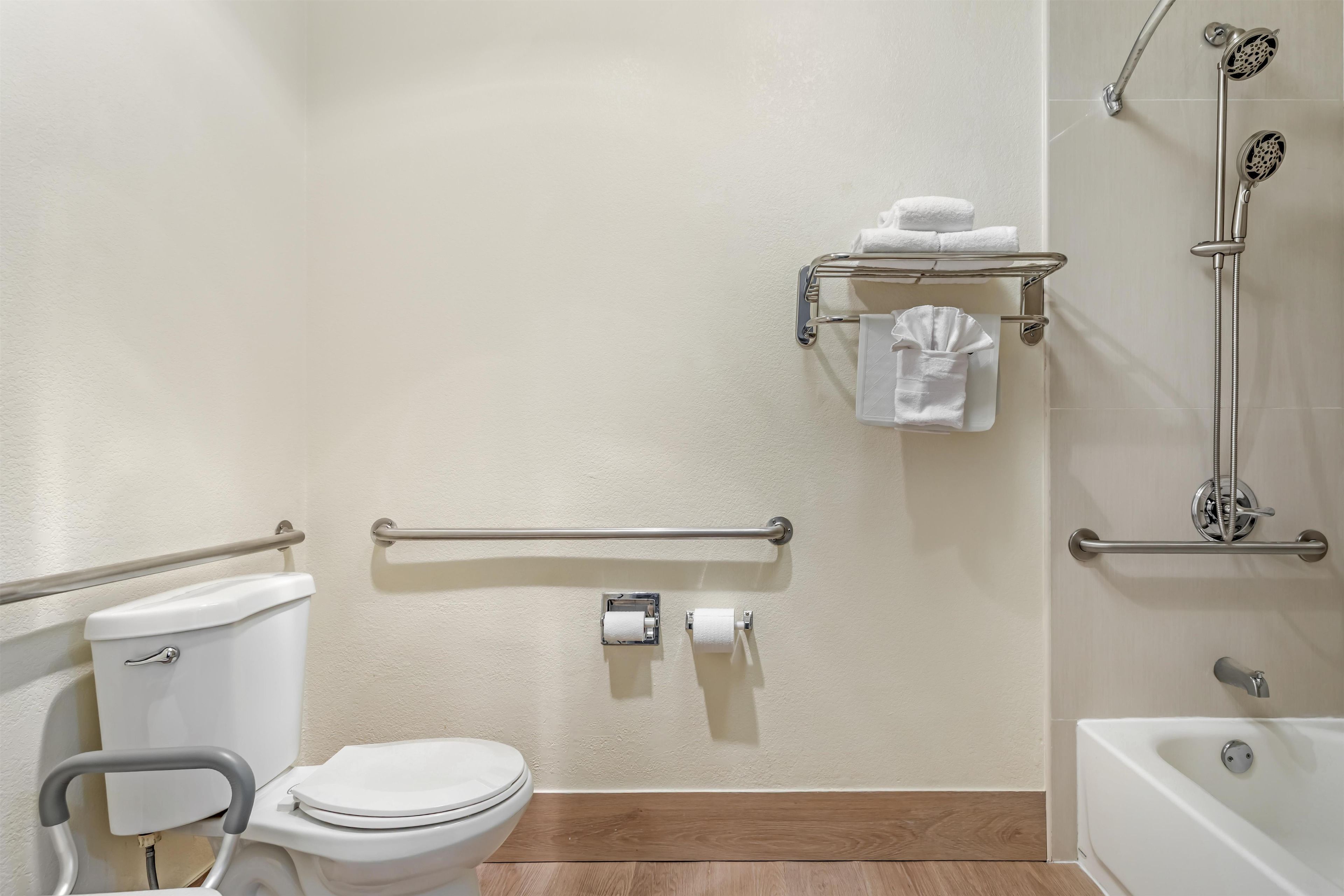 A bathroom with a toilet, a hand grip on the toilet seat, a grab bar, a towel rack with towels, a toilet paper holder, and a shower with a handheld showerhead.