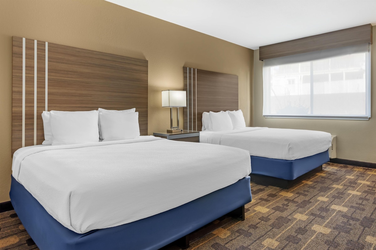 A hotel room with two beds, a nightstand, a lamp, and a large window.