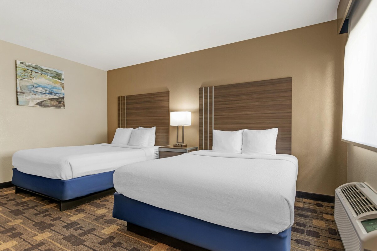 A hotel room with two beds, a nightstand, a lamp, a painting, and a window with blinds.