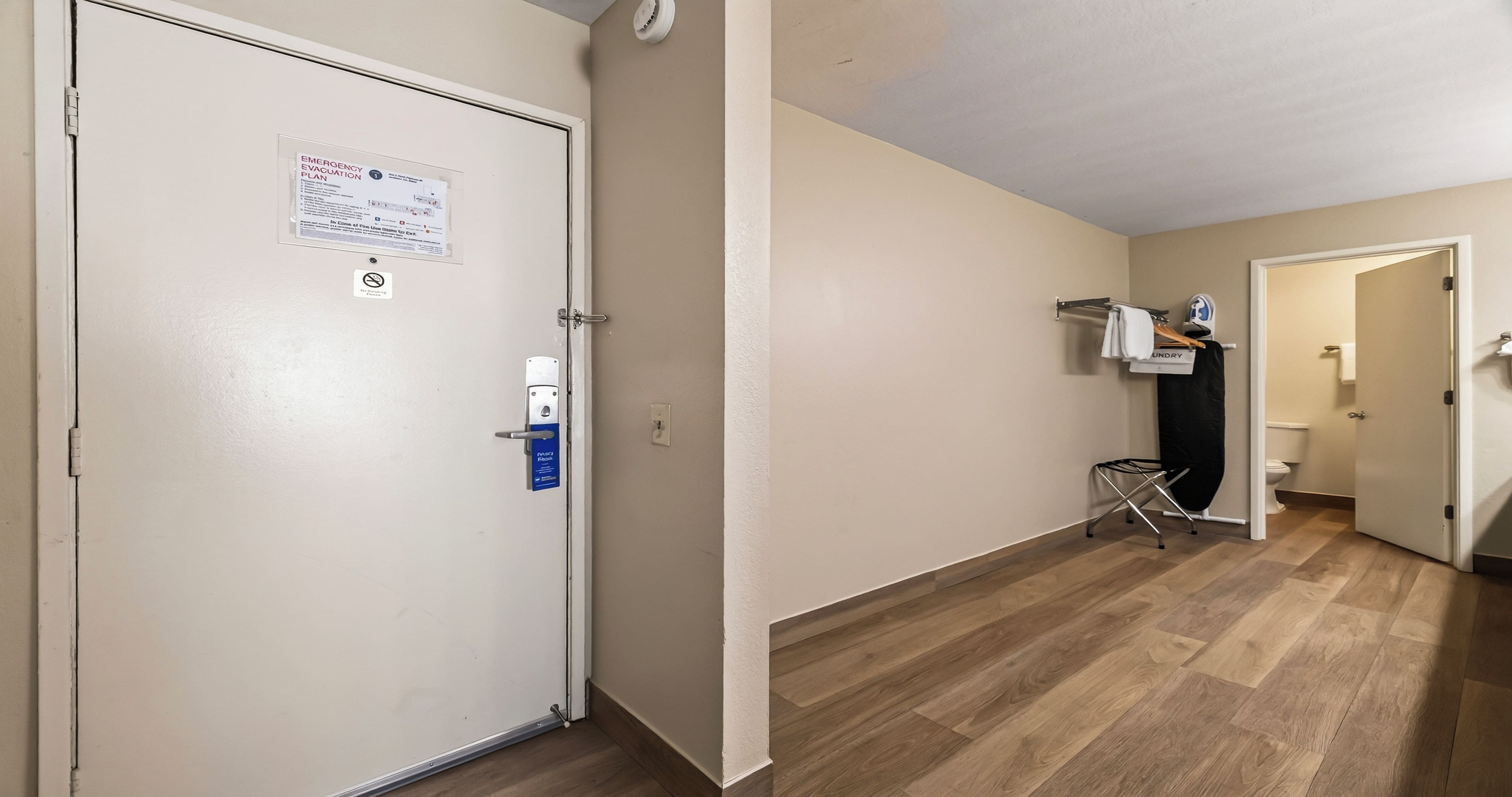 Ein Flur mit einer geschlossenen Tür links, einem an der Wand befestigten Notfallplan, einem Rauchverbotsschild und einem Waschbereich mit Kleiderstange und Bügeleisen.