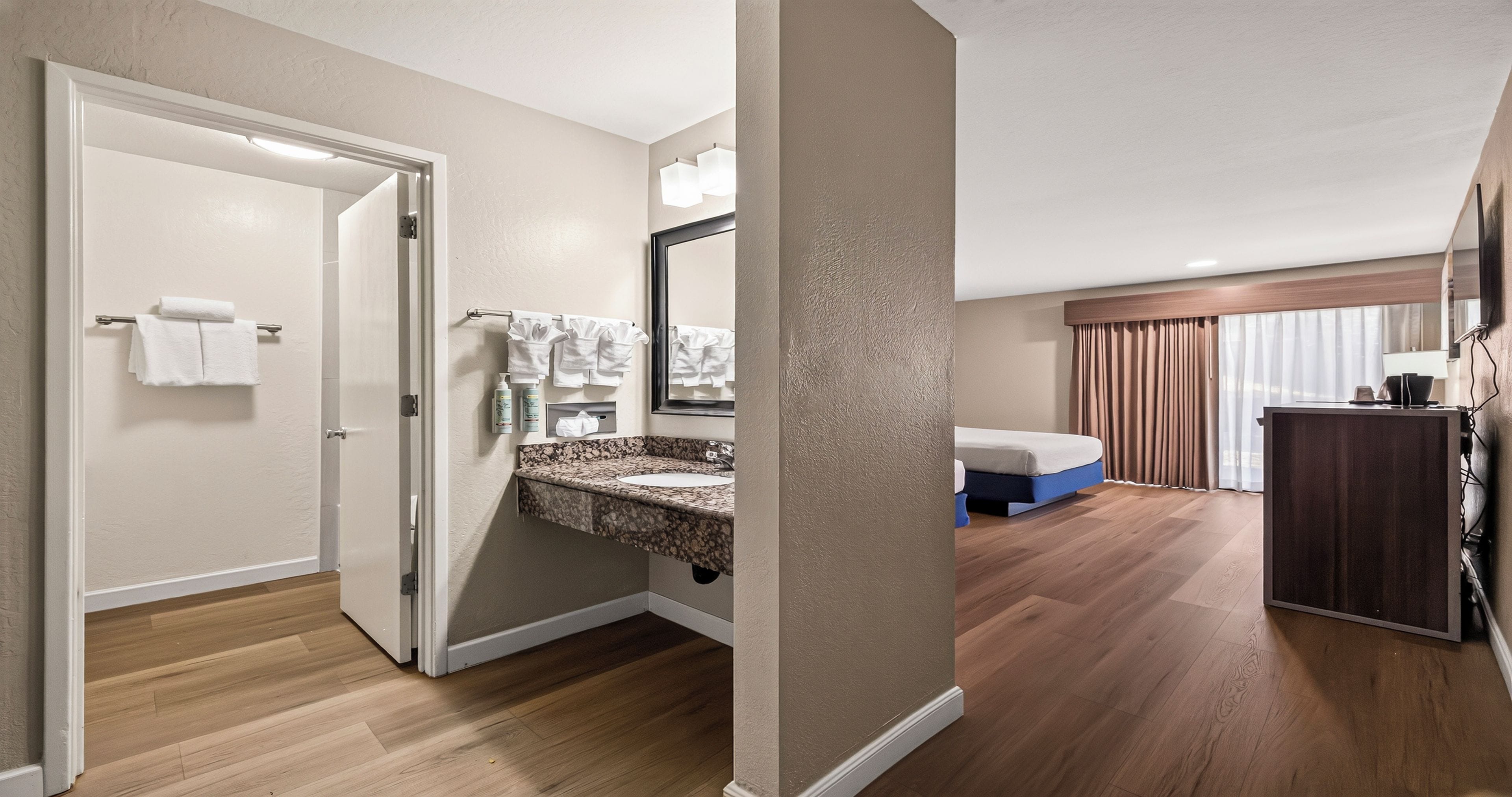 A hotel room with a bathroom, a bed, a desk, and a large window with curtains.