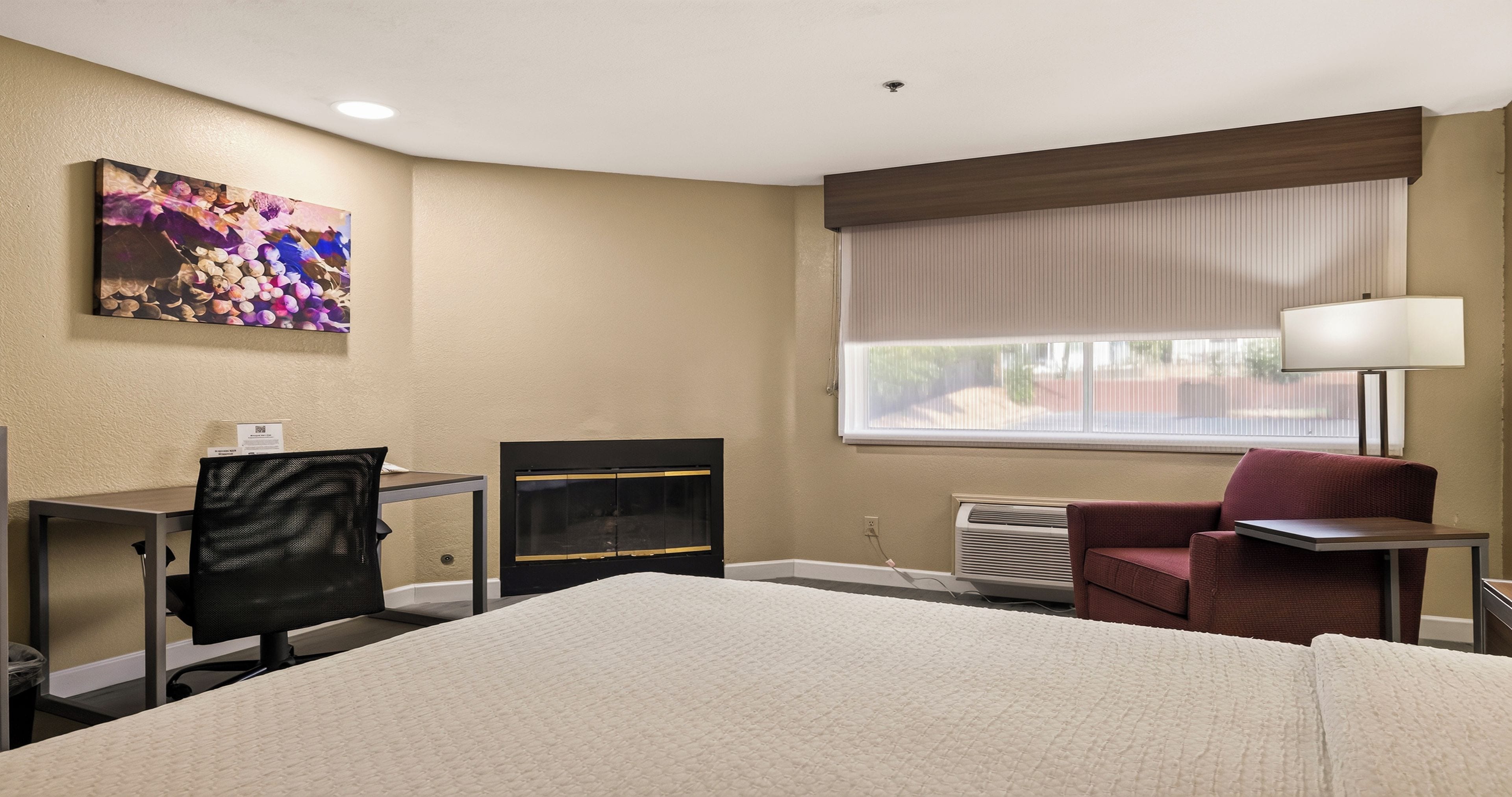 A hotel room with a bed, a desk with a chair, a fireplace, a window with blinds, and a lamp.