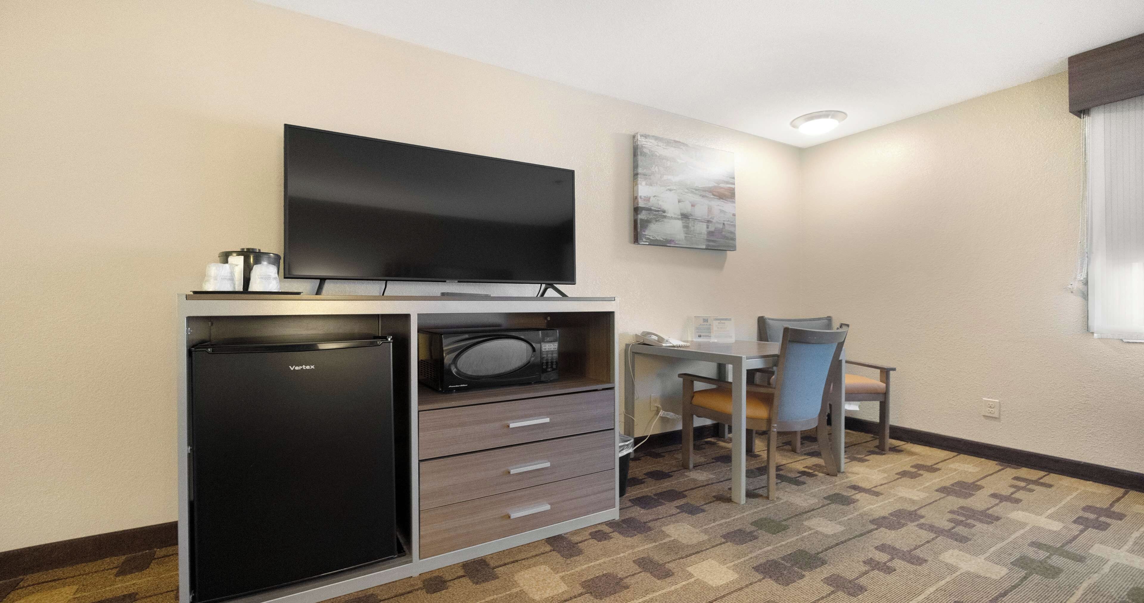A compact hotel room with a flat-screen TV, a refrigerator, a microwave, a small table with a chair, and a painting on the wall.
