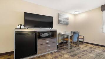 A compact hotel room with a flat-screen TV, a refrigerator, a microwave, a small table with a chair, and a painting on the wall.