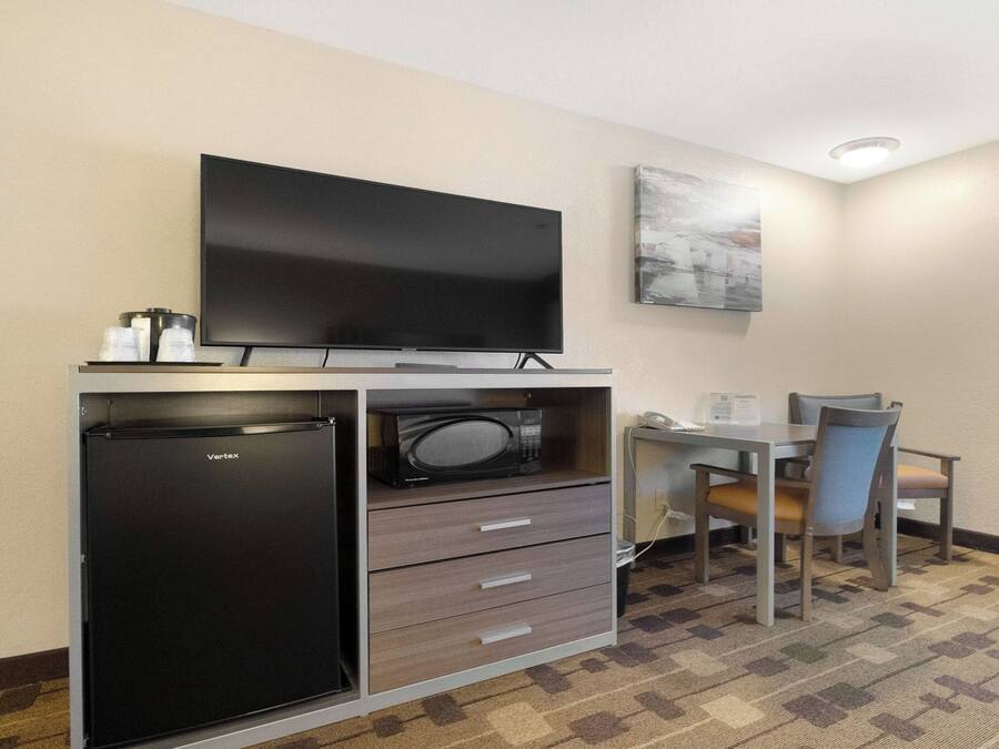 A compact hotel room with a flat-screen TV, a refrigerator, a microwave, a small table with a chair, and a painting on the wall.