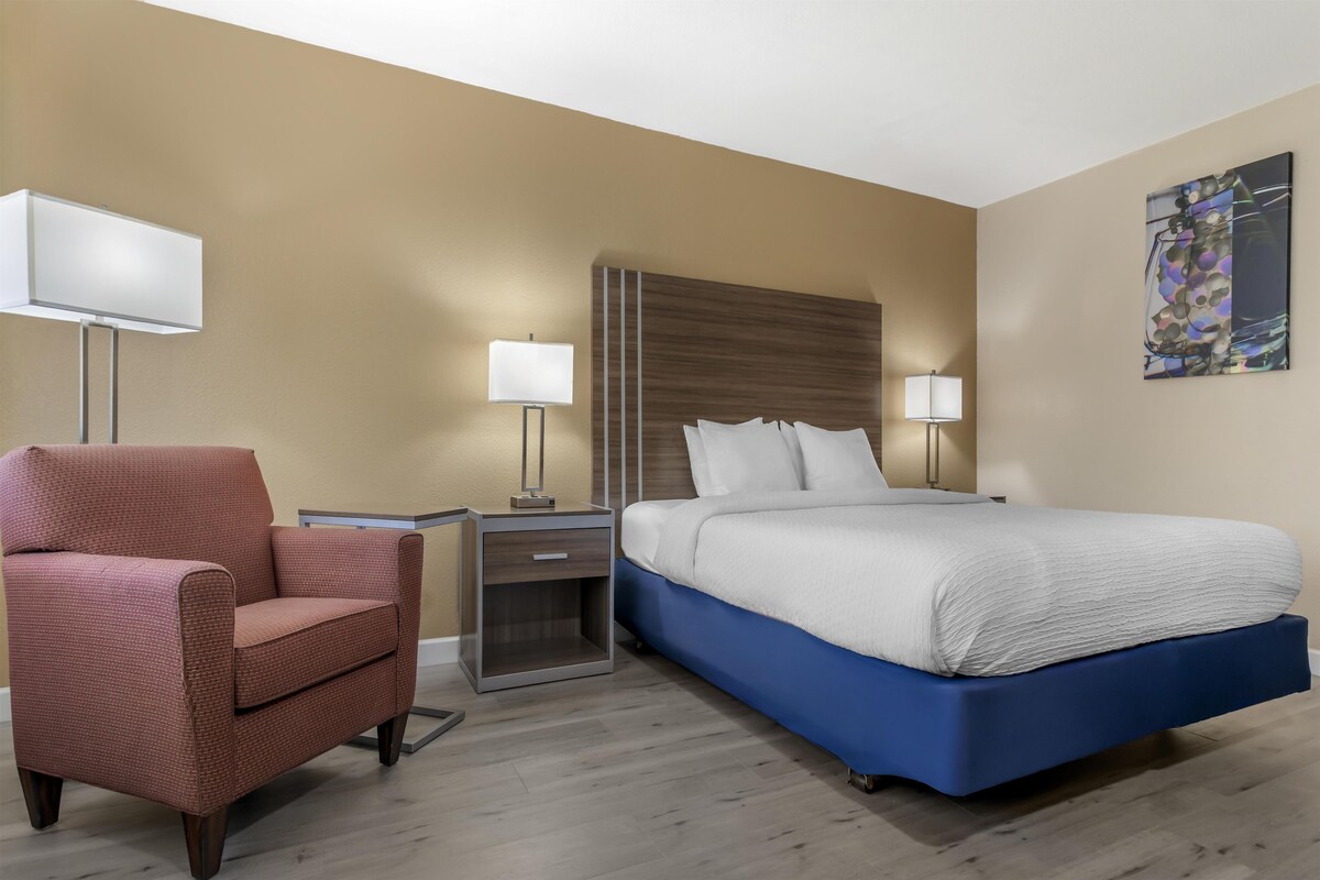 A hotel room with a bed, a chair, a nightstand, a lamp, and a painting on the wall.