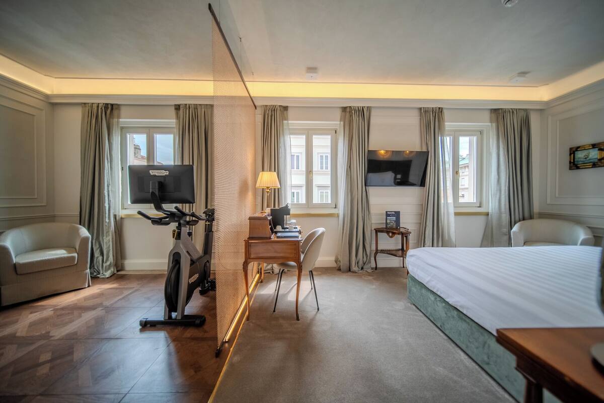 Habitación de hotel con cama, escritorio, silla, bicicleta estática, televisor y ventanas con cortinas.