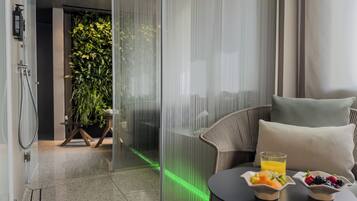 A modern bathroom with a glass shower enclosure, a green plant wall, and a seating area with a table set with fruit and a glass of orange juice.