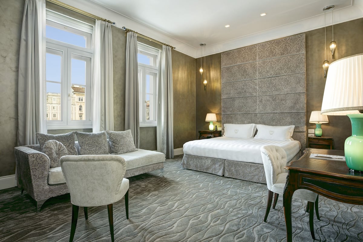 Habitación de hotel con una cama grande, un sofá, una silla, un escritorio y vistas a edificios a través de la ventana.