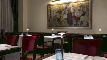 A restaurant with red velvet chairs, white tablecloths, and a large painting on the wall.