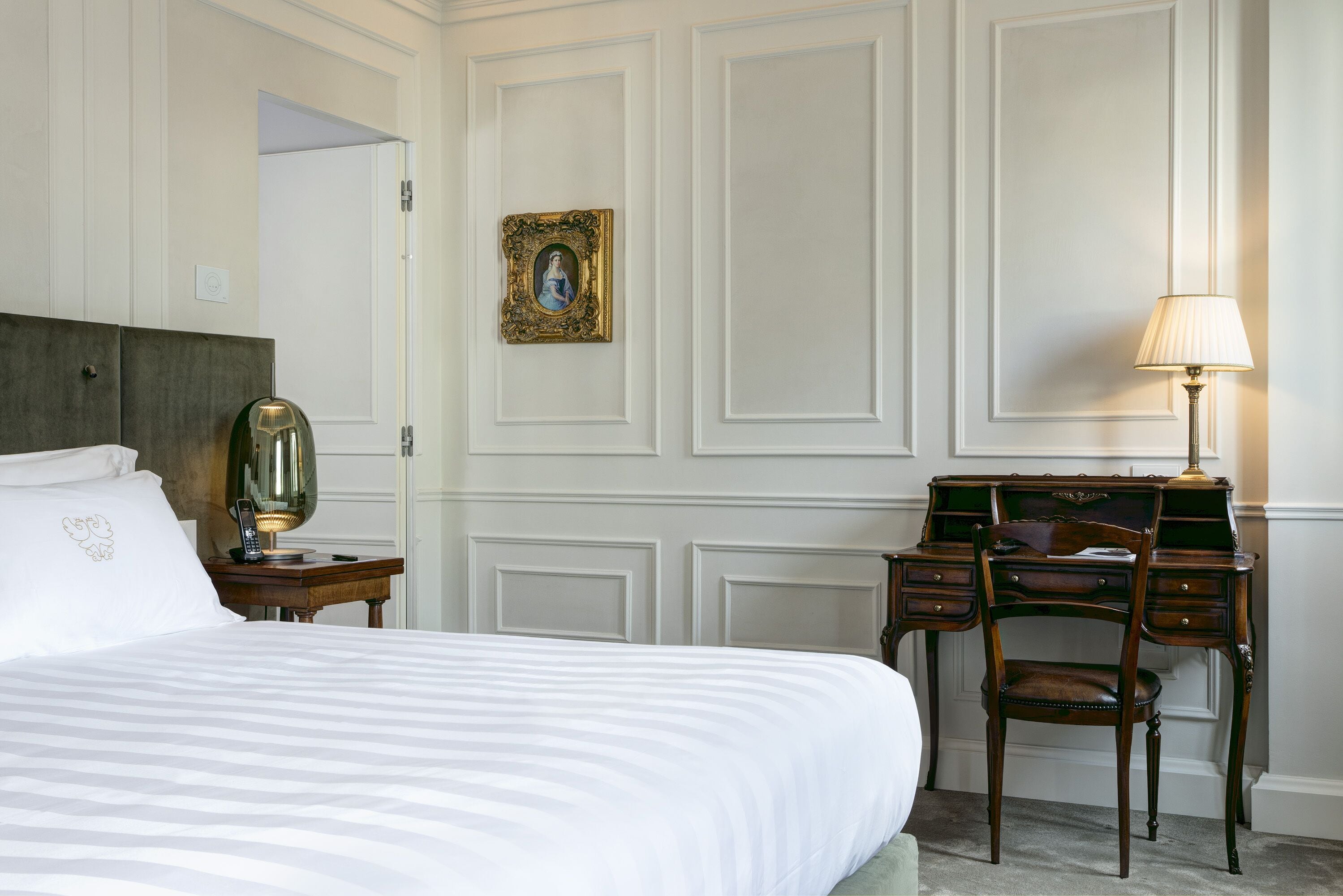 A hotel room with a bed, a desk, a chair, a lamp, and a framed picture on the wall.