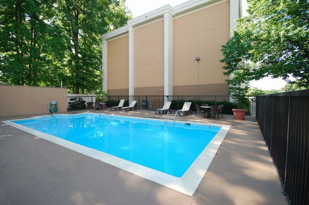 Piscina exterior con sillas de descanso y una mesa, rodeada por una valla y árboles.
