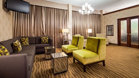 A hotel room with a dark-colored sofa, two green armchairs, a glass coffee table, a lamp, and a television mounted on the wall.