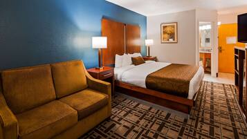 A hotel room with a bed, a sofa, a nightstand, a lamp, a framed picture, and a television.