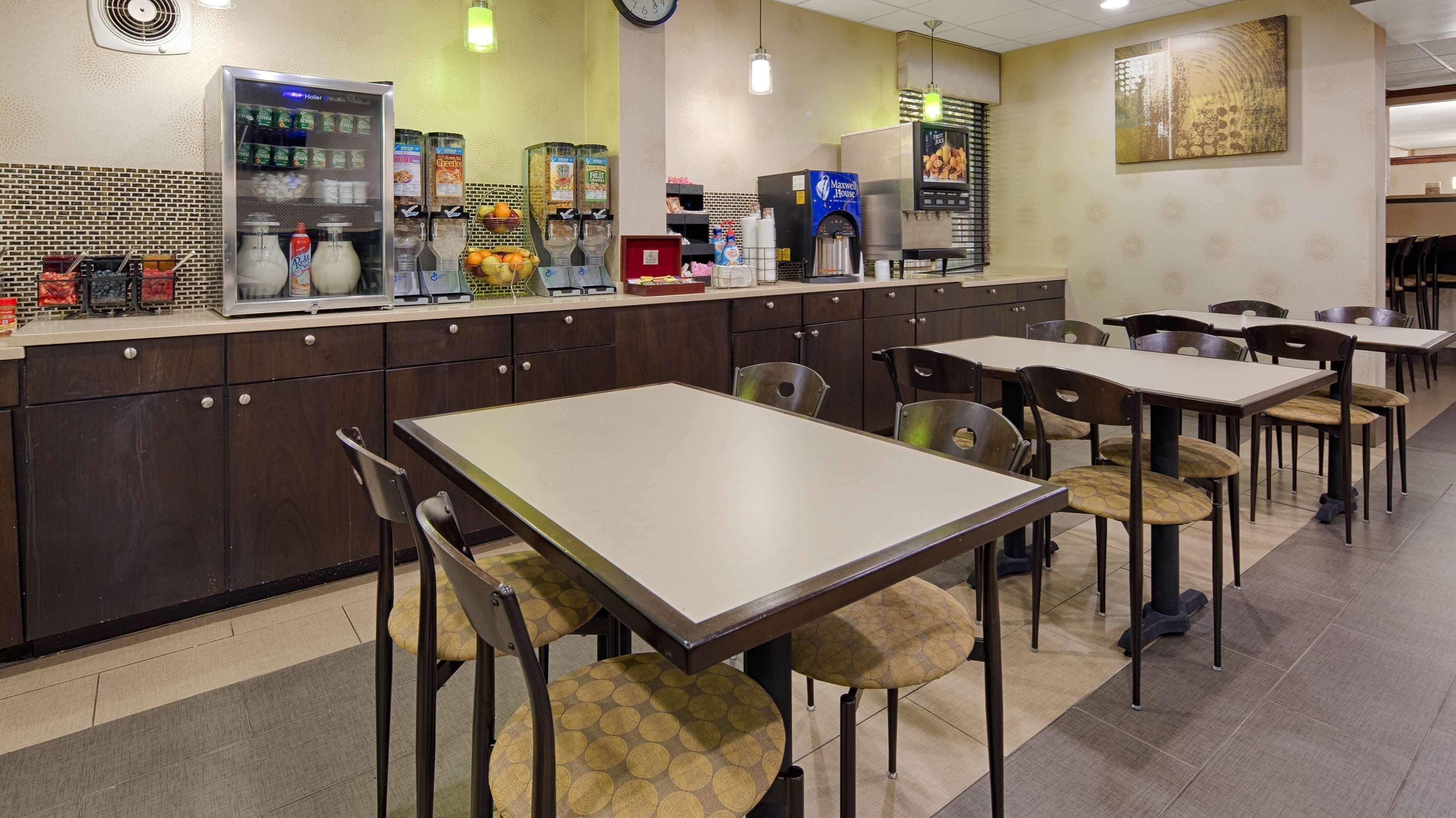 Una cafetería con dispensadores automáticos de bebidas, una máquina de café y una variedad de alimentos en el mostrador. Hay varias mesas y sillas dispuestas para comer.