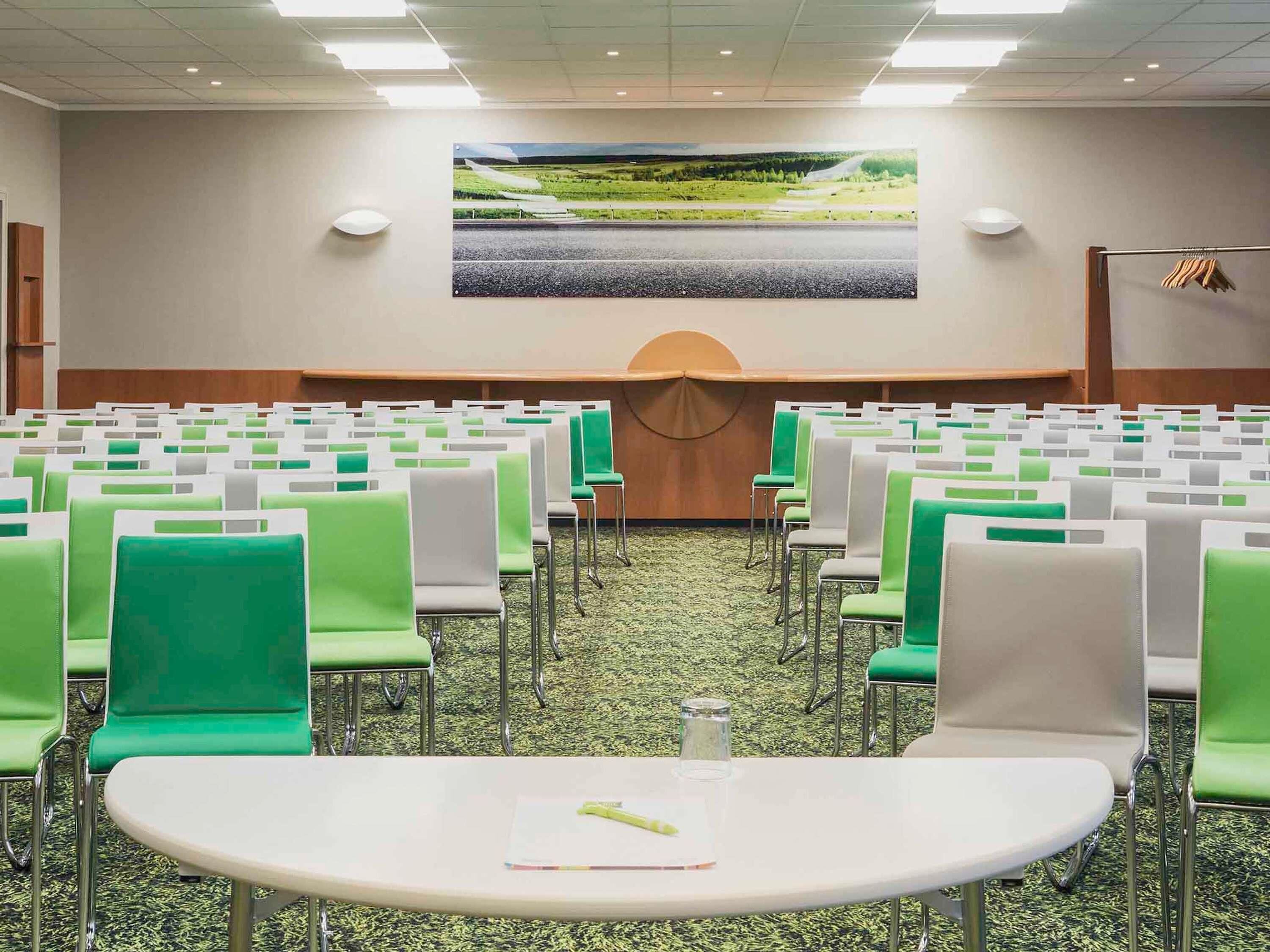Een vergaderzaal met rijen groene en witte stoelen, een groot scherm aan de muur en een vloerbedekking.