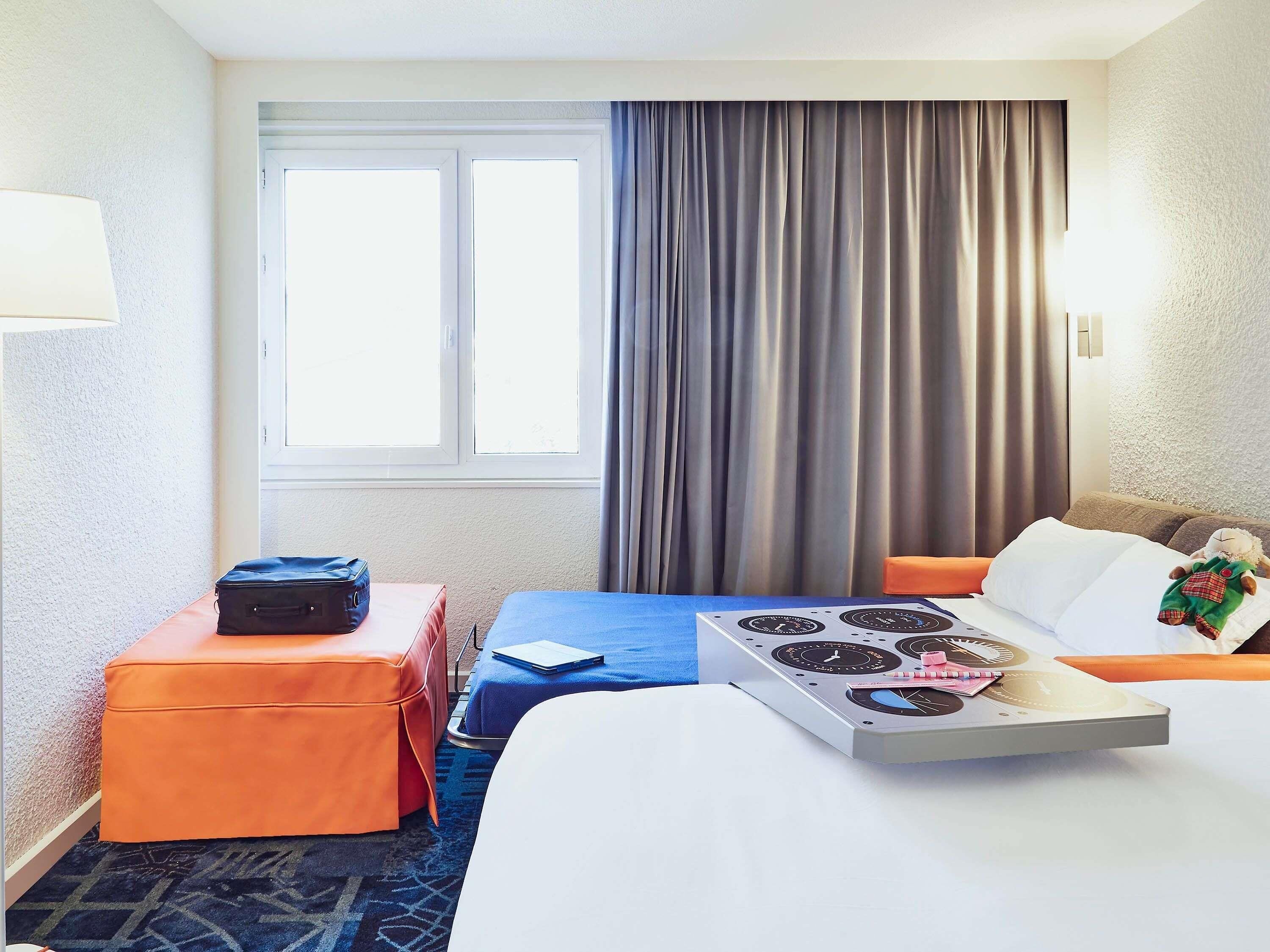 Ein Hotelzimmer mit einem Bett, einem kleinen Tisch mit Wasserkocher, einem Koffer und einem Kuscheltier.