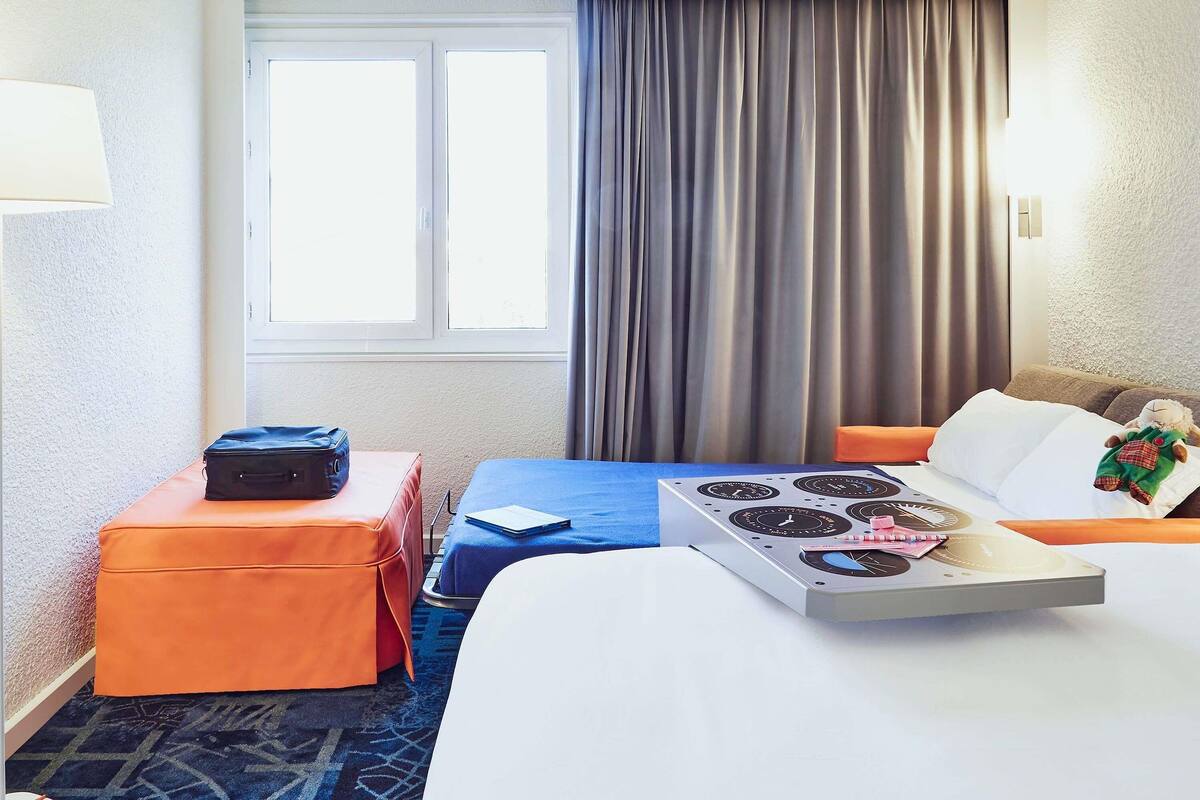Een hotelkamer met een bed, een tafeltje met een waterkoker, een koffer en een pluchen speeltje op het bed.