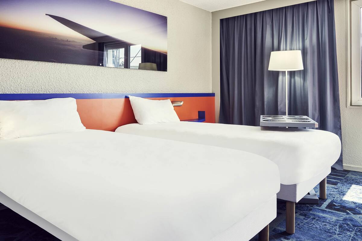 Een hotelkamer met twee bedden, een raam met gordijnen, een lamp en een aan de muur hangend kunstwerk.
