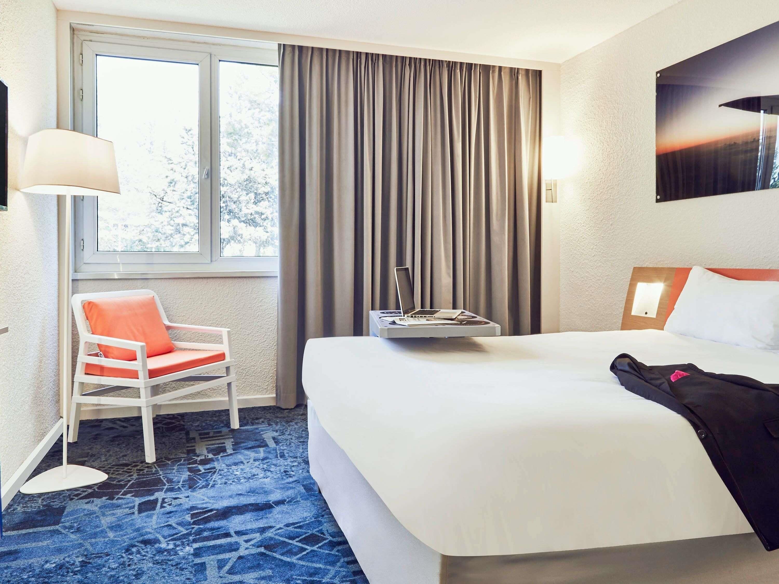 Ein Hotelzimmer mit einem Bett, einem Sessel, einem Schreibtisch mit einem Laptop, einer Lampe und einem Fenster mit Vorhängen.