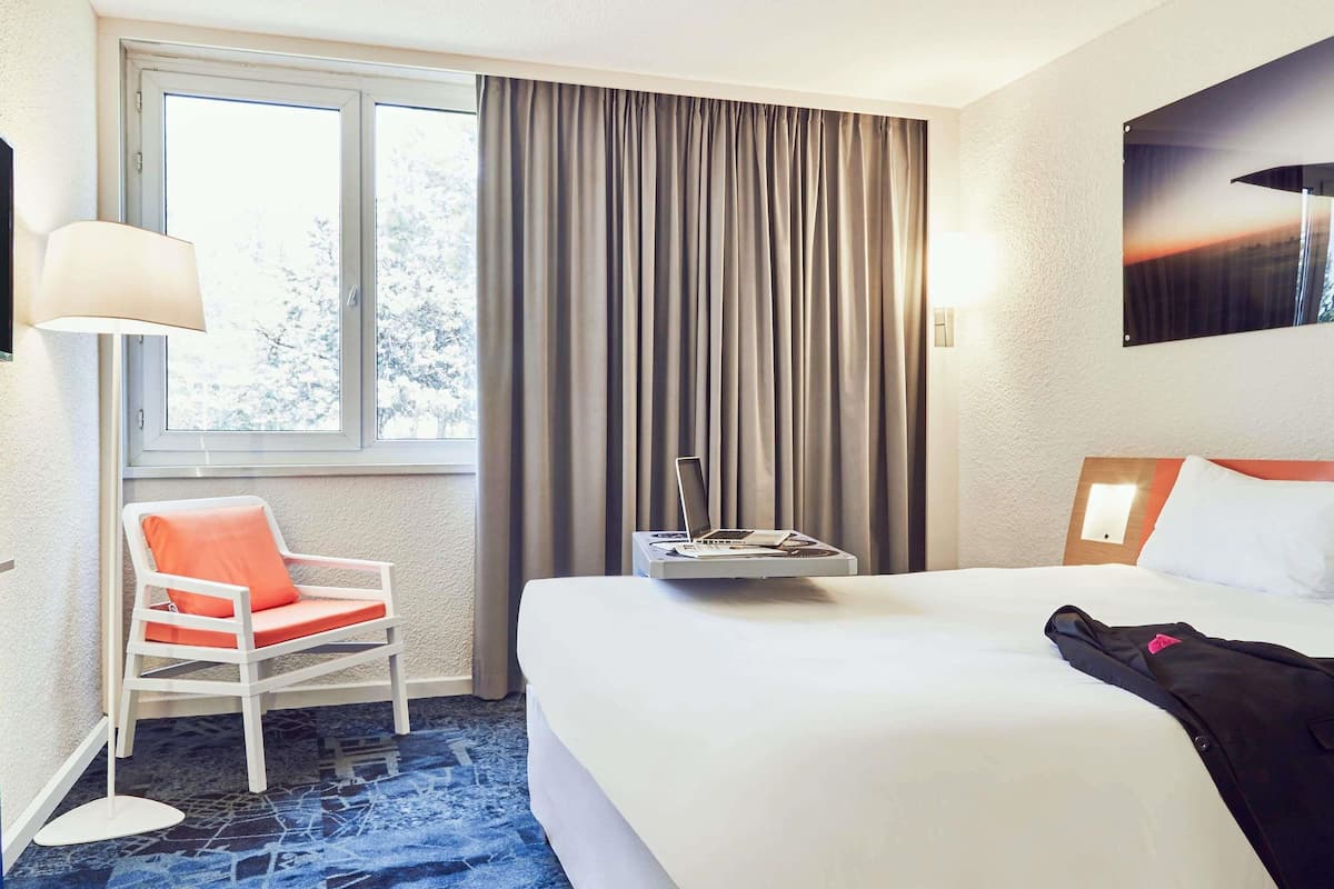 Een hotelkamer met een bed, een stoel, een bureau met een laptop, een raam met gordijnen en een wandlamp.