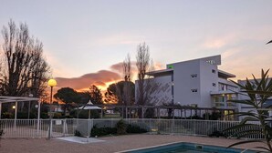 Un hotel moderno con piscina, rodeado por una valla blanca, y una puesta de sol al fondo.
