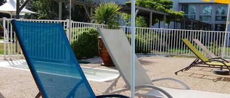 Outdoor lounge chairs with umbrellas, a white fence, and a building in the background.