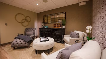Una habitación bien iluminada con un sillón capitonado, un otoman blanco y un sofá con cojines decorativos. Un mueble negro con un espejo encima está apoyado contra la pared.