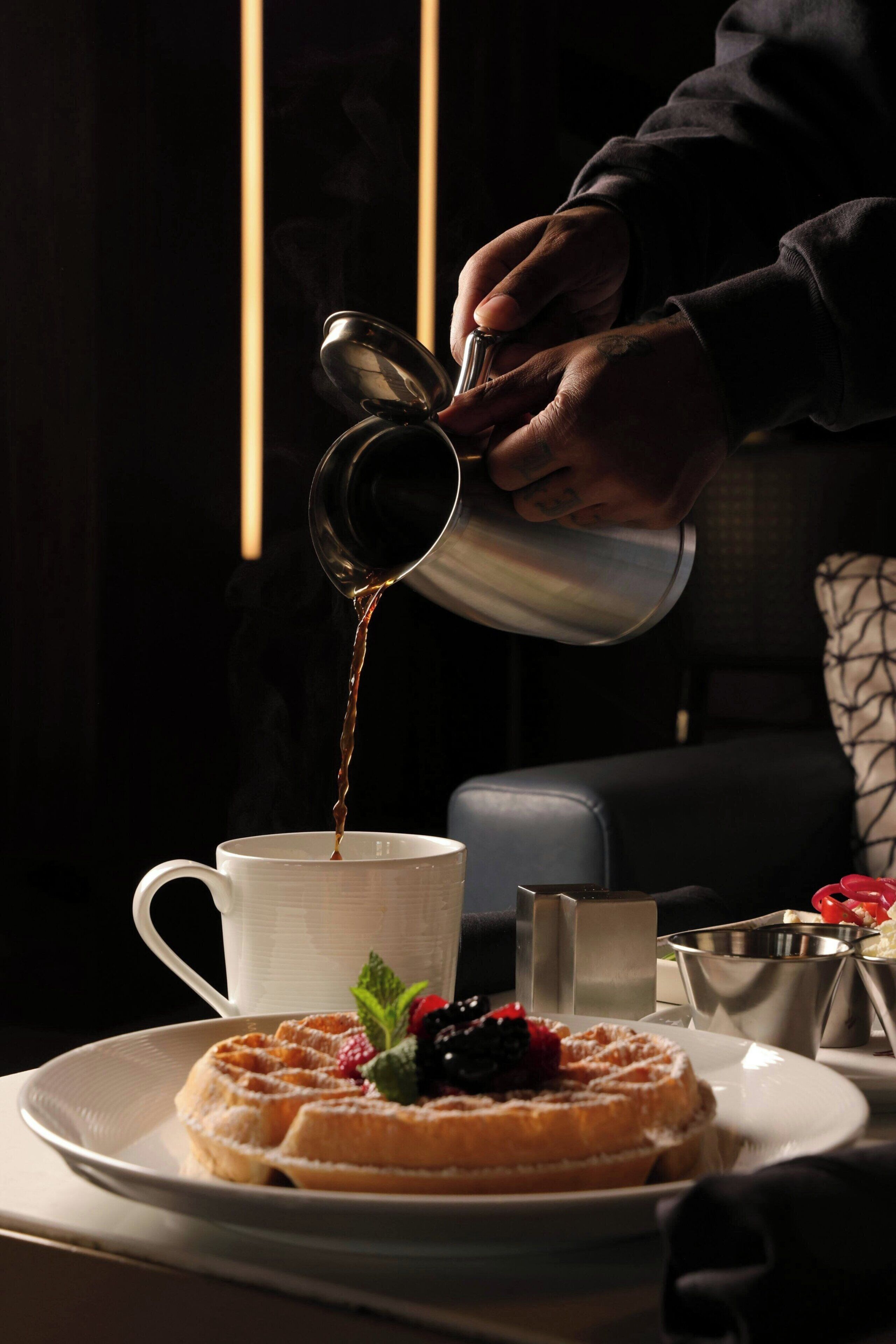 A person pouring a beverage into a cup, a plate with a dessert, and a small metal container on a table.