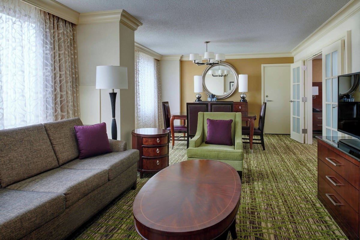 A hotel room with a sofa, armchair, dining table, and a TV.