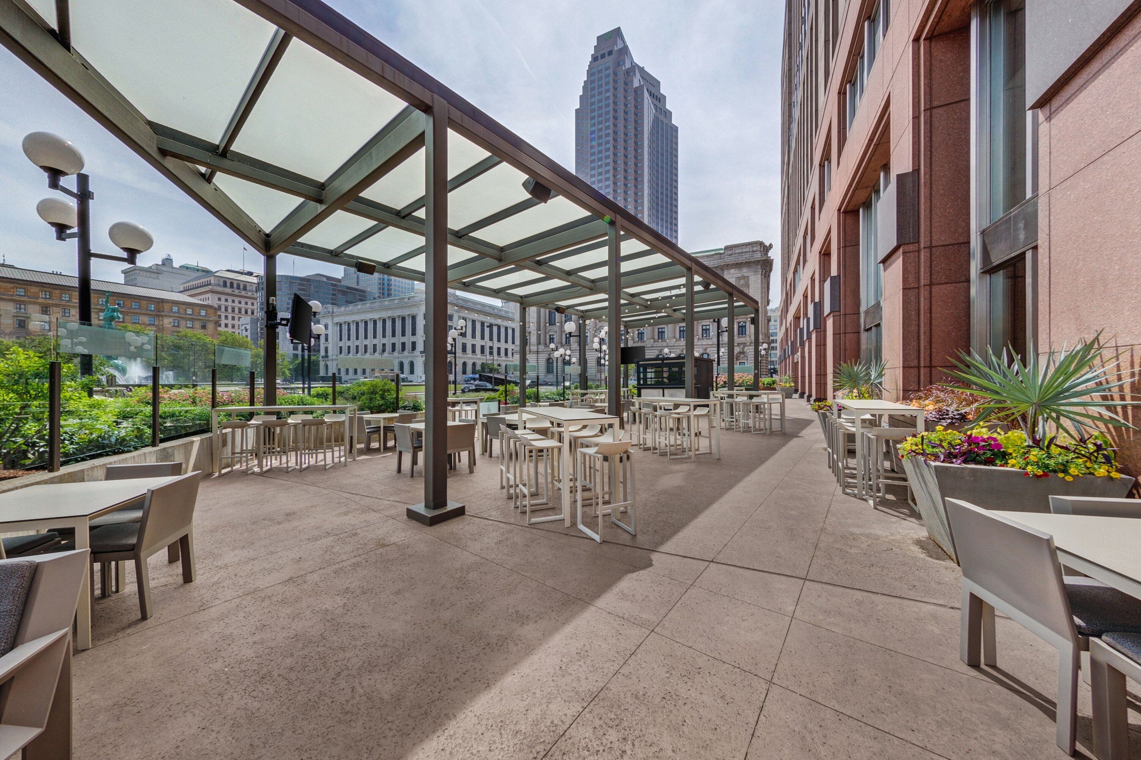 An outdoor café with tables and chairs, a glass roof, and a cityscape view.