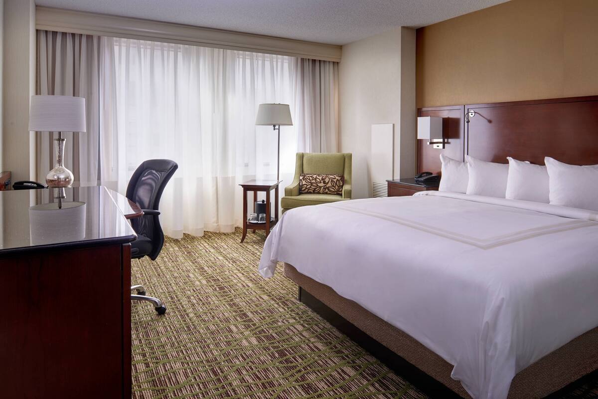 A hotel room with a large bed, a desk with a lamp, a chair, a small table, and a nightstand.