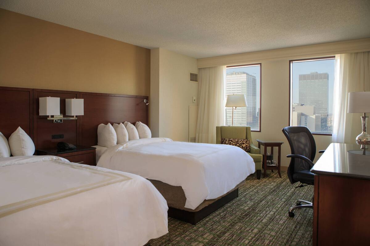 A hotel room with two beds, a desk, a chair, a lamp, and a view of the city through large windows.