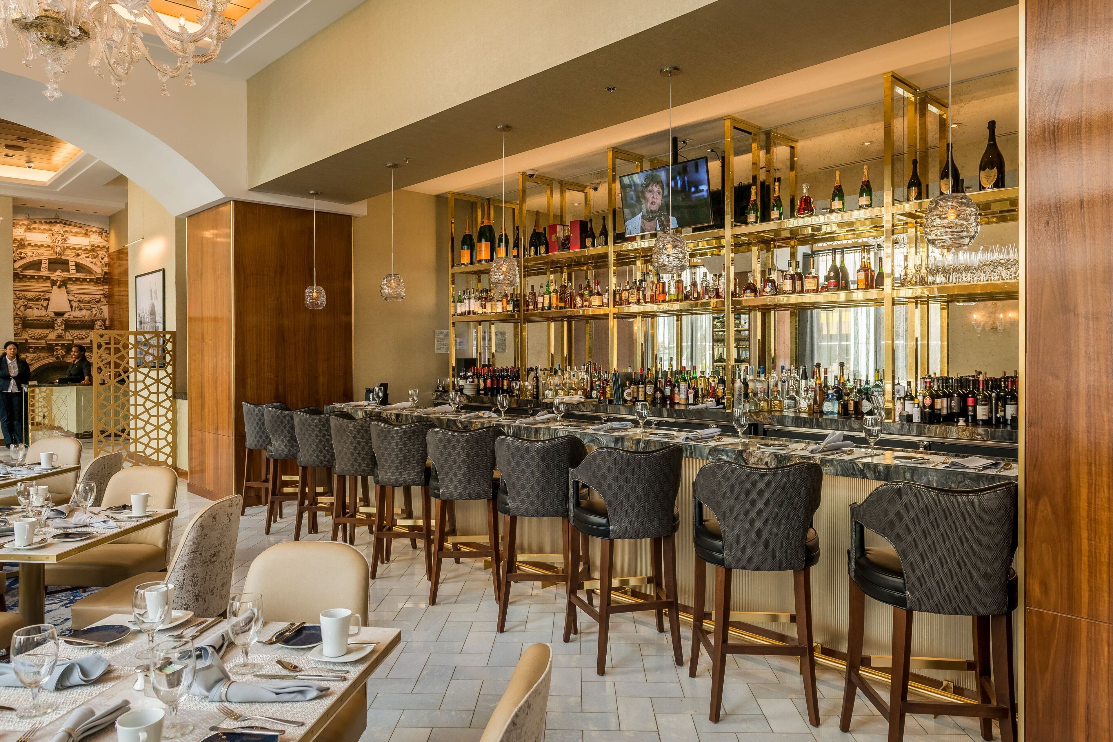 A bar area with multiple high stools, a television mounted on the wall, and shelves stocked with bottles and glasses.