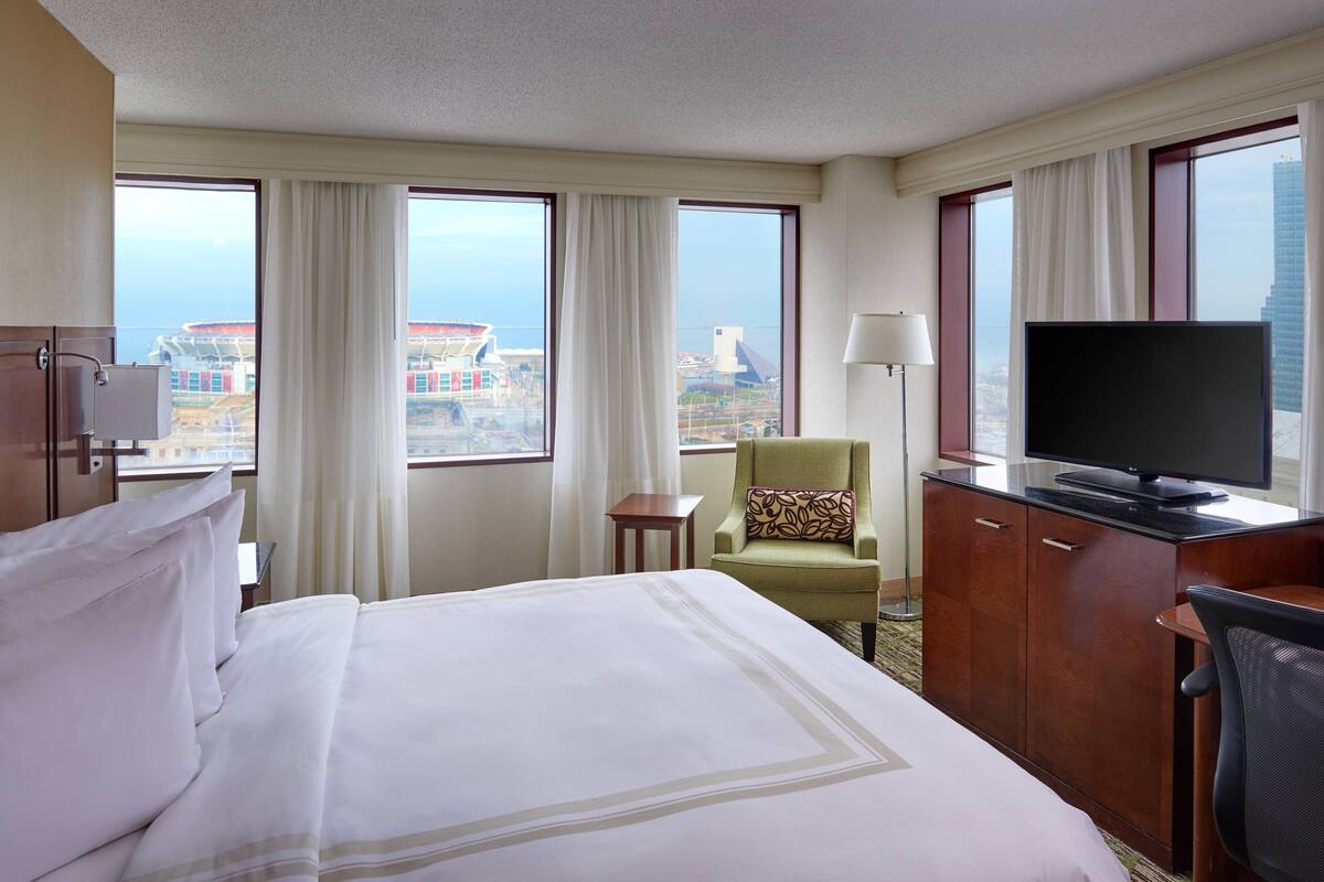 A hotel room with a bed, a desk with a TV, a chair, and large windows offering a city view.