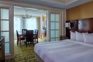 A hotel room with a large bed, a desk with a chair, and a view through an open door to a dining area.
