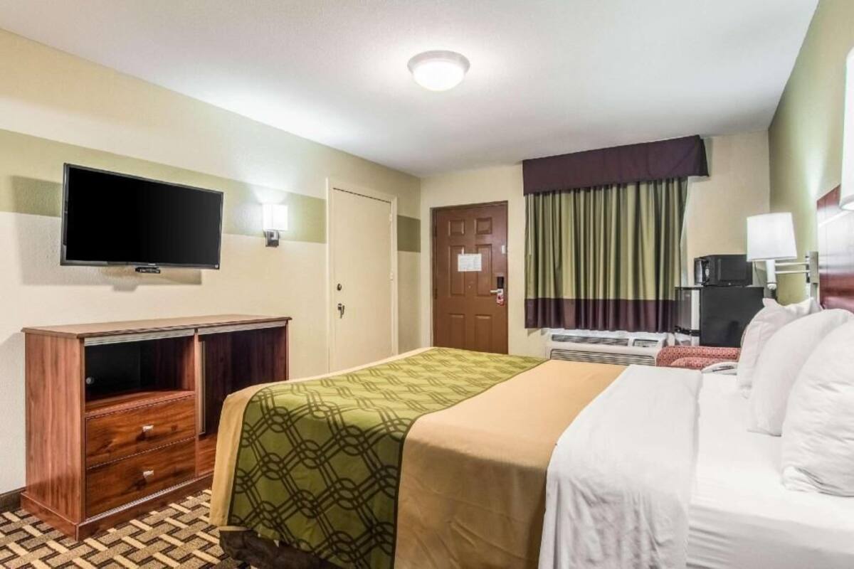 Quarto de hotel com cama, televisão, escrivaninha e janela com cortinas.