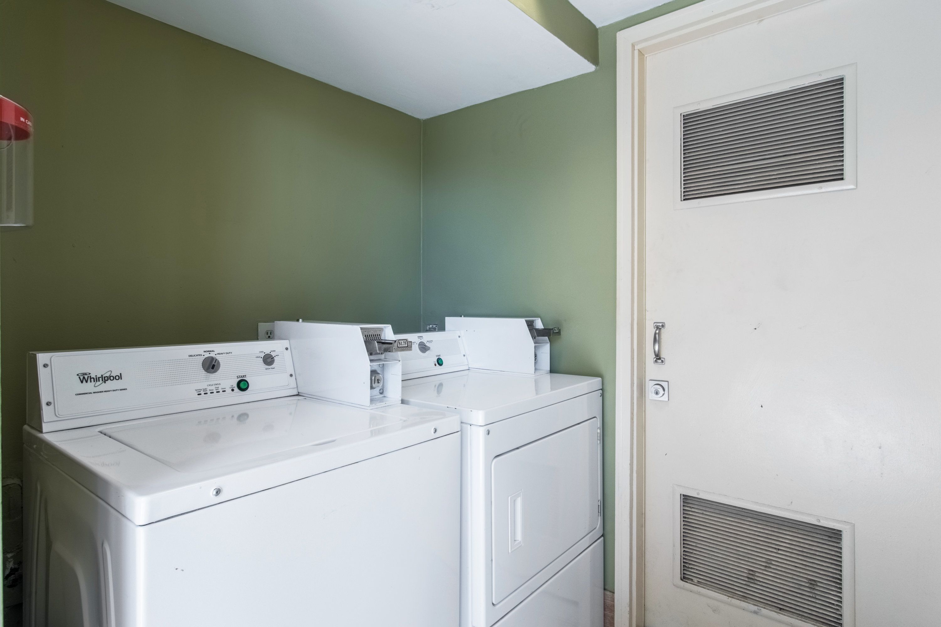 Un área de lavandería con dos lavadoras y secadoras apiladas, una pared verde y una puerta con rejilla de ventilación.